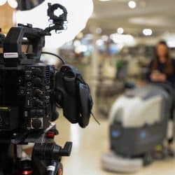 Detailed side view of a Canon cinema camera on a tripod in a retail store. The background is blurred, showing a woman standing behind a large commercial floor cleaner.
