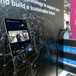 Tablet on a flexible stand displaying a promotional video of a cleaning equipment demonstration, set against a black wall with a global network graphic and text promoting sustainable technology.