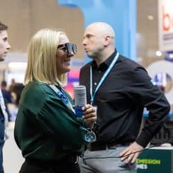 Smiling woman wearing smart glasses interacts with a mounted tablet at an exhibition booth. She holds a can of sparkling water while two men with event lanyards stand nearby, observing.