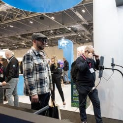 Busy trade show floor with two men at the front testing smart glasses connected to mounted tablets. Other attendees and branded booths, including "ICE" and "bcc", are visible in the background.
