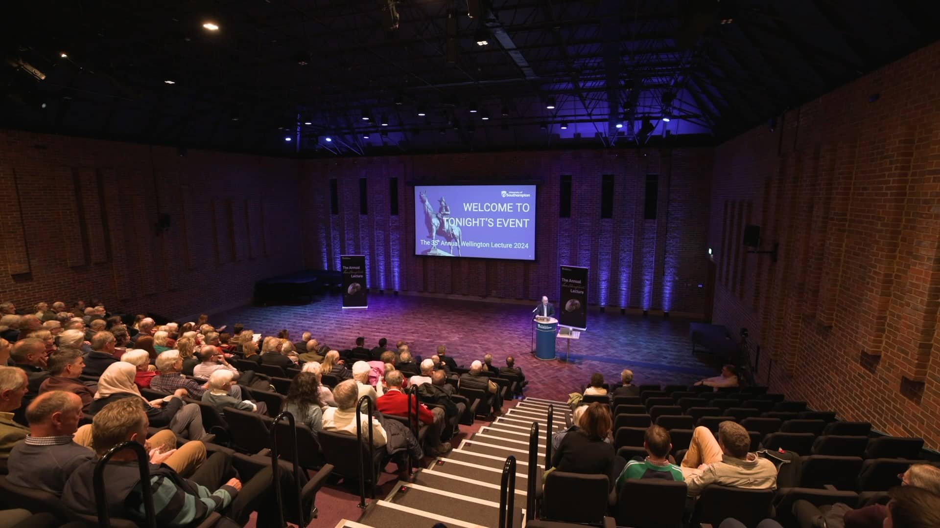 A wide view of a packed auditorium at the Turner Sims concert hall during the 35th Annual Wellington Lecture 2024. A speaker stands at a podium on stage in front of a large screen displaying the event title, while the audience listens attentively. The venue features exposed brick walls, purple stage lighting, and tiered seating.