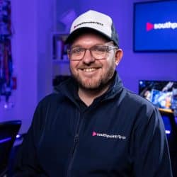 A person with short brown hair, a beard, and glasses wearing a navy blue jacket and a baseball cap with the southpoint films logo. They are standing in a video production studio with blue ambient lighting and computer monitors in the background.