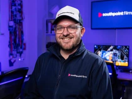 A person with short brown hair, a beard, and glasses wearing a navy blue jacket and a baseball cap with the southpoint films logo. They are standing in a video production studio with blue ambient lighting and computer monitors in the background.