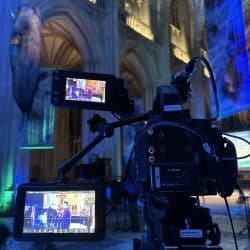 A close-up, low-angle shot of a professional video camera on a tripod, positioned in a large cathedral. The camera has two attached monitors, both displaying a live feed of people on a stage. In the background, the intricate stone arches of the cathedral are visible, illuminated by blue and green lights, and a large blue whale-like structure hangs from the ceiling.