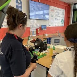 A videographer wearing a Southpoint Films polo shirt films a cooking activity involving children in a classroom kitchen. The scene includes a countertop with chopped vegetables, a white mixing bowl, and instructional slides displayed on a screen in the background.