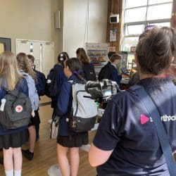 A group of secondary school students in uniforms queue up in a school cafeteria. A woman wearing a "Southpoint Films" shirt stands in the foreground, filming the scene with a professional camera. Bright daylight comes through large windows in the background.