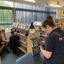 Inside a school library, a woman from "Southpoint Films" films students who are browsing books and DVDs on low shelves. Three students sit and kneel near a display, engaged in discussion. The room is well-lit with natural light from large vertical windows.