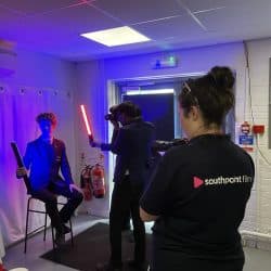 A student in a blazer poses with two LED light wands in a photography studio setup. Another student photographs him while a woman from "Southpoint Films" records the session with a video camera. Coloured lighting creates a dramatic blue and red effect.