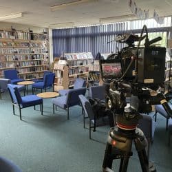 Inside a brightly lit library, a large black professional video camera mounted on a red and black tripod is the central focus. The camera faces towards the left of the frame, capturing a scene with multiple bookshelves filled with books, several small round tables with blue chairs around them, and a window with blue blinds in the background. A colorful, inflatable figure stands near the bookshelves on the left.