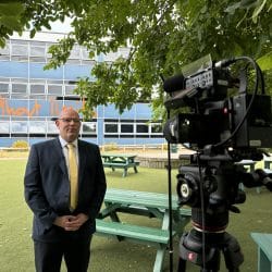A man in a suit and yellow tie stands outdoors in front of a school building, giving an interview. A professional video camera on a tripod is set up in the foreground, with picnic benches and a leafy tree framing the scene.
