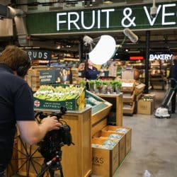 A film crew is setting up to record in the produce section of a grocery store. A cameraman wearing a "southpoint films" shirt operates a professional video camera on a tripod, pointed towards a man standing near displays of fruits and vegetables under a "FRUIT & VEG" sign. He is operating a floor cleaning machine. A boom microphone is positioned overhead, held by another crew member who can be seen in the background near the bakery section.