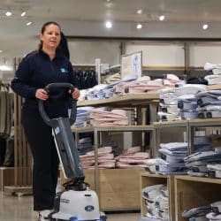 A woman in a dark blue long-sleeved shirt and dark trousers is operating a white and grey floor cleaning machine in a retail clothing store. She is standing and looking towards the right side of the frame. Behind her are displays of folded shirts on shelves and racks of hanging clothes. The lighting is bright, typical of a retail environment.