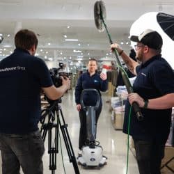 A film crew of four is recording a scene in a retail clothing store. A woman in a dark blue sweater and black pants stands facing the camera, holding a white object. A cameraman behind a tripod-mounted camera and a sound operator with a boom microphone face her. Racks of clothing are visible in the background.