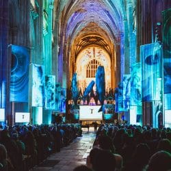 An interior shot of a large, old cathedral with rows of people seated, facing a stage at the far end of the hall. Tall, blue and green banners depicting abstract, organic shapes hang from the high arched ceiling. Two large, dark blue whale-like structures hang near the stage. Two screens on either side of the hall, closer to the viewer, display images of whales. The overall lighting is dim, with blue and green spotlights illuminating the banners and stage.