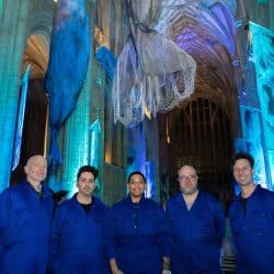 Five people, four men and one woman, stand in a line facing the camera, all wearing matching royal blue jumpsuits. They are in a large, old cathedral with high vaulted ceilings and large, ornate windows in the background. Several large, dark blue whale-like structures and translucent, net-like installations hang from the ceiling. The cathedral is illuminated with blue and purple lighting.