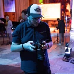 A man wearing a dark blue polo shirt, black-rimmed glasses, and a white and black "Vans Off The Wall" trucker hat is looking down and adjusting a black camera with a large lens. He is standing in a cathedral, illuminated by purple and blue lighting, with blurred figures and architectural details in the background.