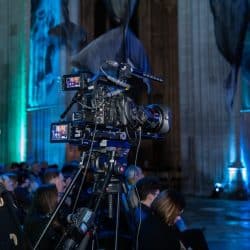 A medium shot from behind a woman with long, brown hair and headphones, looking up and to the right. In front of her, a professional video camera on a tripod is prominently featured, with its monitors displaying a live feed. In the background, rows of people are seated in a dimly lit cathedral, with large banners hanging from the ceiling, illuminated by blue and green lights.
