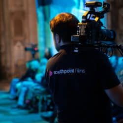 A view from behind a person wearing a dark polo shirt with "southpoint films" written on the back, holding a professional camera on their shoulder. They are facing away from the viewer, looking towards a dimly lit space in a cathedral where people are seated. Blue and green lighting illuminates the scene.