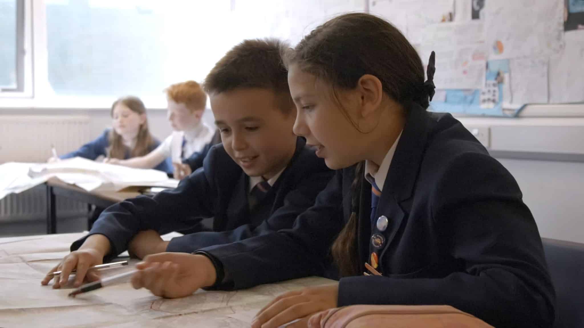 Two young students wearing dark blue school blazers, white shirts, and ties are closely collaborating on a document spread out on a desk. The student on the right, a girl with dark hair pulled back, is speaking while gesturing at the paper. The student on the left, a boy with short brown hair, is looking down at the paper and pointing. Other students are visible working at desks in the blurred background of the classroom.