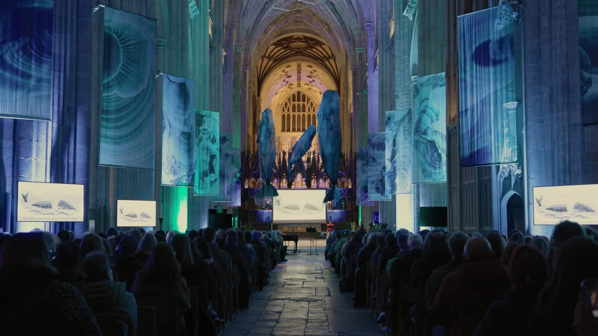 An interior shot of a large, old cathedral with rows of people seated, facing a stage at the far end of the hall. Tall, blue and green banners depicting abstract, organic shapes hang from the high arched ceiling. Three large, dark blue whale-like structures hang near the stage. Two screens on either side of the hall, closer to the viewer, display images of whales. The overall lighting is dim, with blue and green spotlights illuminating the banners and stage.