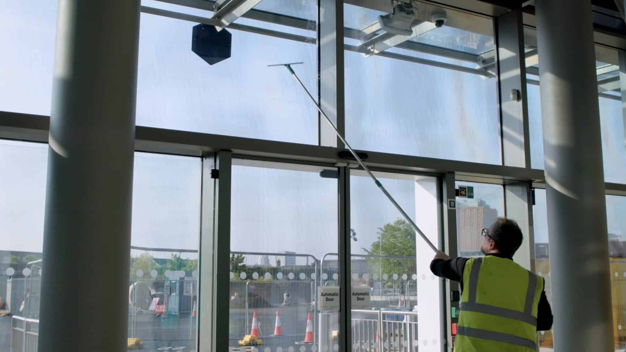 A worker in a high-visibility vest cleaning tall glass windows inside a modern building using an extended cleaning pole, with daylight streaming through the glass panels.