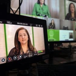A close-up shot of a small video monitor displaying a woman speaking, with an on-screen timecode of "00:00:00:00". In the blurred background, a larger monitor shows a grid of multiple video conference participants