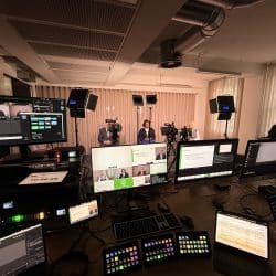 A wide shot of a live production control room setup. In the foreground, a large table is filled with multiple computer monitors, laptops, keyboards, and control panels. Various video feeds and a countdown timer are visible on the screens. In the background, three people are seated and being filmed on a small stage with professional lighting.