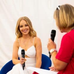 A smiling woman with long blonde hair, wearing a white sleeveless dress, is seated and holding a microphone. She is looking towards another person, seen from behind, who is also holding a microphone and appears to be interviewing her. They are seated in blue chairs against a white backdrop, possibly inside a tent or temporary structure.