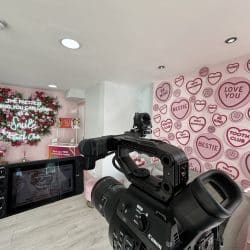 A close-up view from behind a professional video camera, which is pointing towards a pink-walled dental reception area. A large, heart-shaped floral display with neon text "THE PRETTIEST THING YOU CAN WEAR IS A SMILE by Tooth Club" is prominent on the back wall, which is also adorned with various pink heart-shaped plaques.