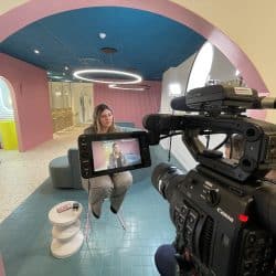 A professional video camera with an external monitor, showing a woman being interviewed, is positioned in the foreground. The background is a stylish dental office interior with pink and blue architectural features, curved seating, and modern lighting.