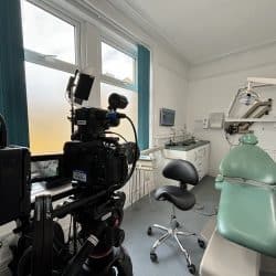 A professional video camera on a tripod is set up inside a dental treatment room. The camera is facing a dental chair and other equipment. Large windows with teal blinds are visible in the background, showing an outdoor view.