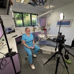 A wide shot inside a bright, modern dental office. A female dental professional in light blue scrubs is seated and smiling at the camera, which is set up on a tripod with a light to the right. Another light stand is visible on the left. Dental equipment, a chair, and a large window looking out onto greenery are in the background.
