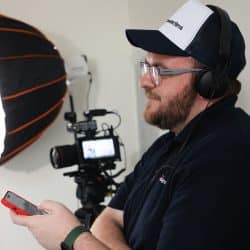 A person wearing a cap and headphones, and a dark polo shirt with the "southpoint films" logo, is looking to the right and holding a red smartphone. A professional video camera setup with a large softbox light is visible in the background on the left.