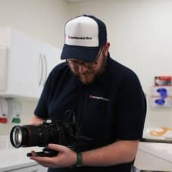 A person wearing a white and blue "southpoint films" cap and a dark polo shirt with the same logo is looking down and adjusting a professional video camera with a large lens. They are standing in what appears to be a dental treatment room with white cabinets and a sink.