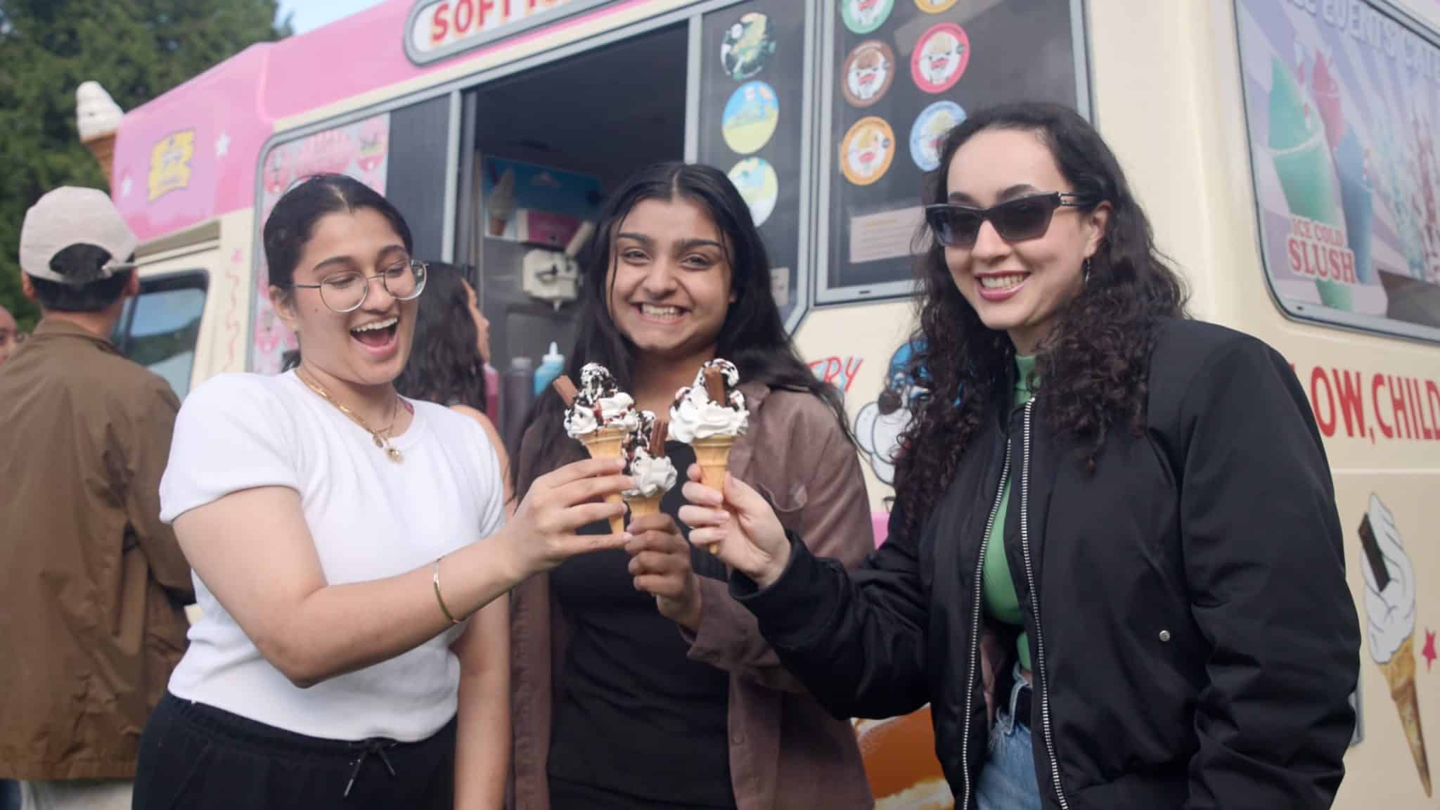 Three smiling women holding ice cream cones and posing in front of a colorful ice cream van, enjoying a cheerful moment at an outdoor event