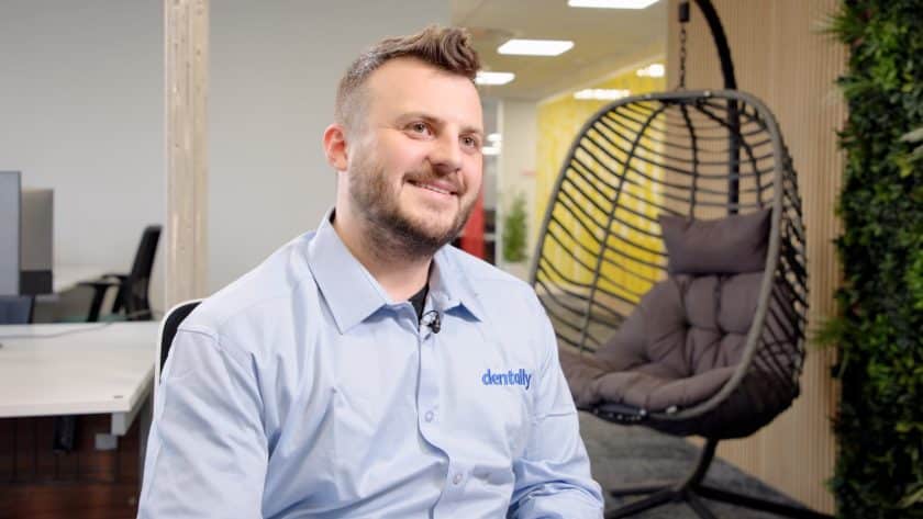 Man smiling during an on-camera interview, wearing a light blue shirt with a 'dentally' logo, seated in a modern office space with a hanging chair and greenery in the background.