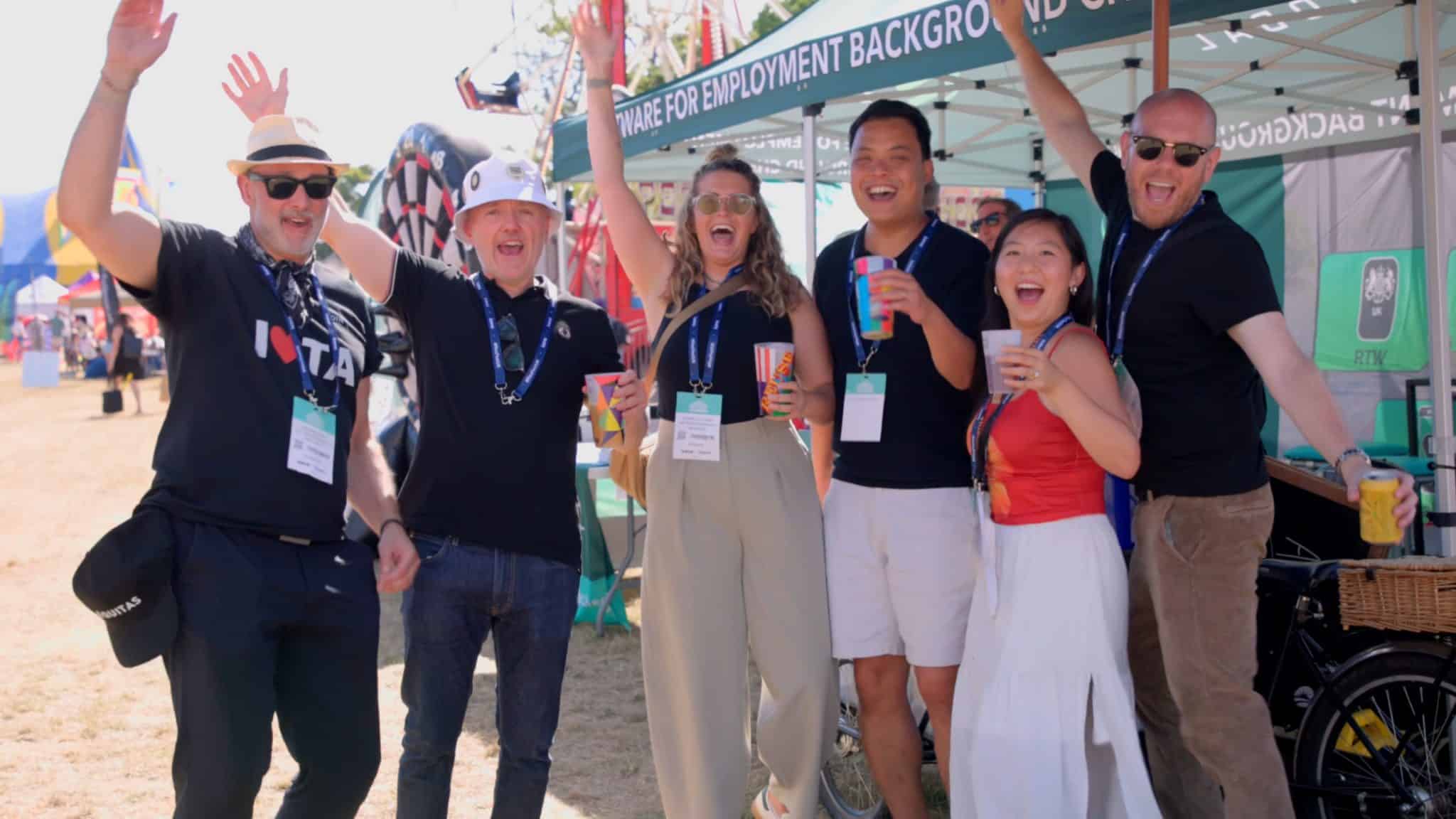 A group of five adults are standing outdoors at what appears to be a festival or event, cheering with their arms raised. They are smiling and holding drinks. In the background, there are various tents and structures. One person on the left wears a hat and sunglasses, and another in the middle wears a white hat.