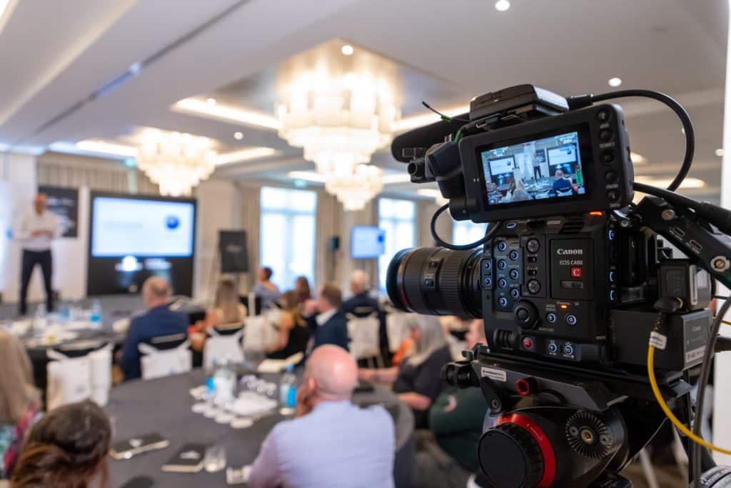 A professional video camera on a tripod in the foreground captures a person giving a presentation in a brightly lit ballroom filled with an audience seated at round tables. The camera's digital screen displays the live feed of the speaker standing next to a large projection screen.