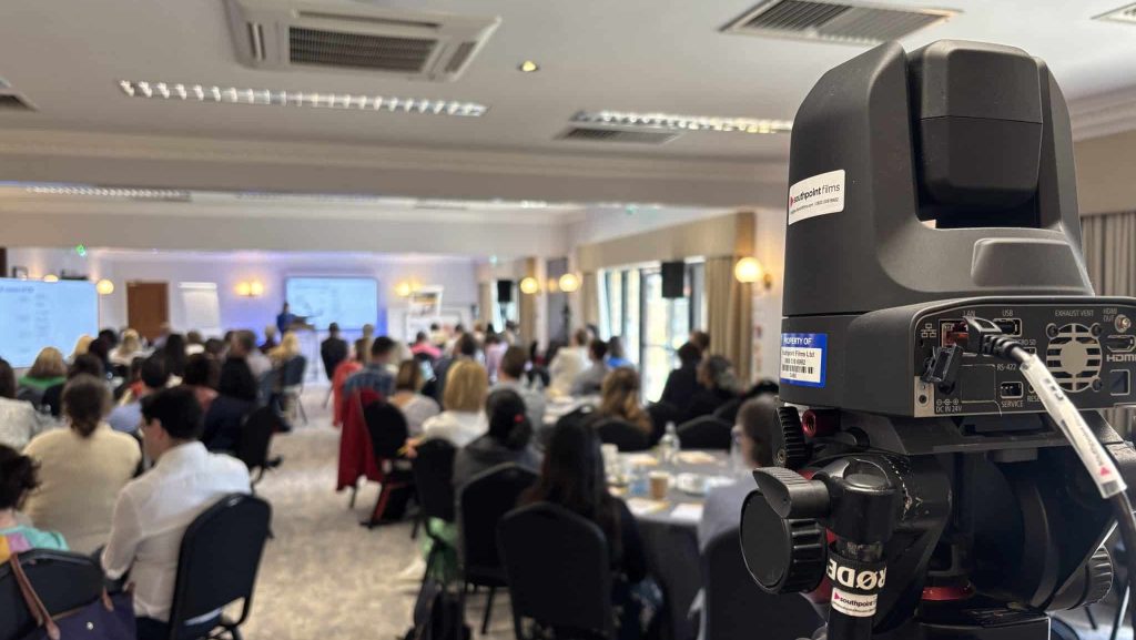 A low-angle, out-of-focus shot of a professional PTZ camera on a tripod in the foreground. In the background, a conference room is filled with people seated at round tables, attending a presentation. A screen is visible at the front of the room.