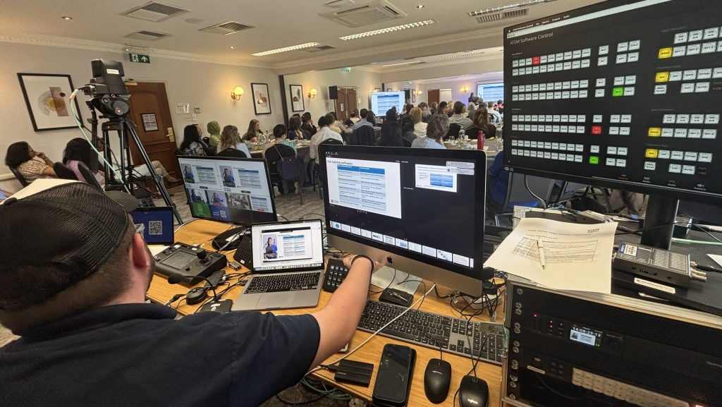 A behind-the-scenes shot of a live event production setup in a conference room. In the foreground, a person wearing a cap is seated at a control desk with multiple computer screens displaying various feeds and controls, including a large monitor with a grid of buttons. In the background, a large audience is seated in rows, watching a presentation on a screen at the front of the room. A professional video camera on a tripod is also visible on the left side of the frame, pointed towards the audience.