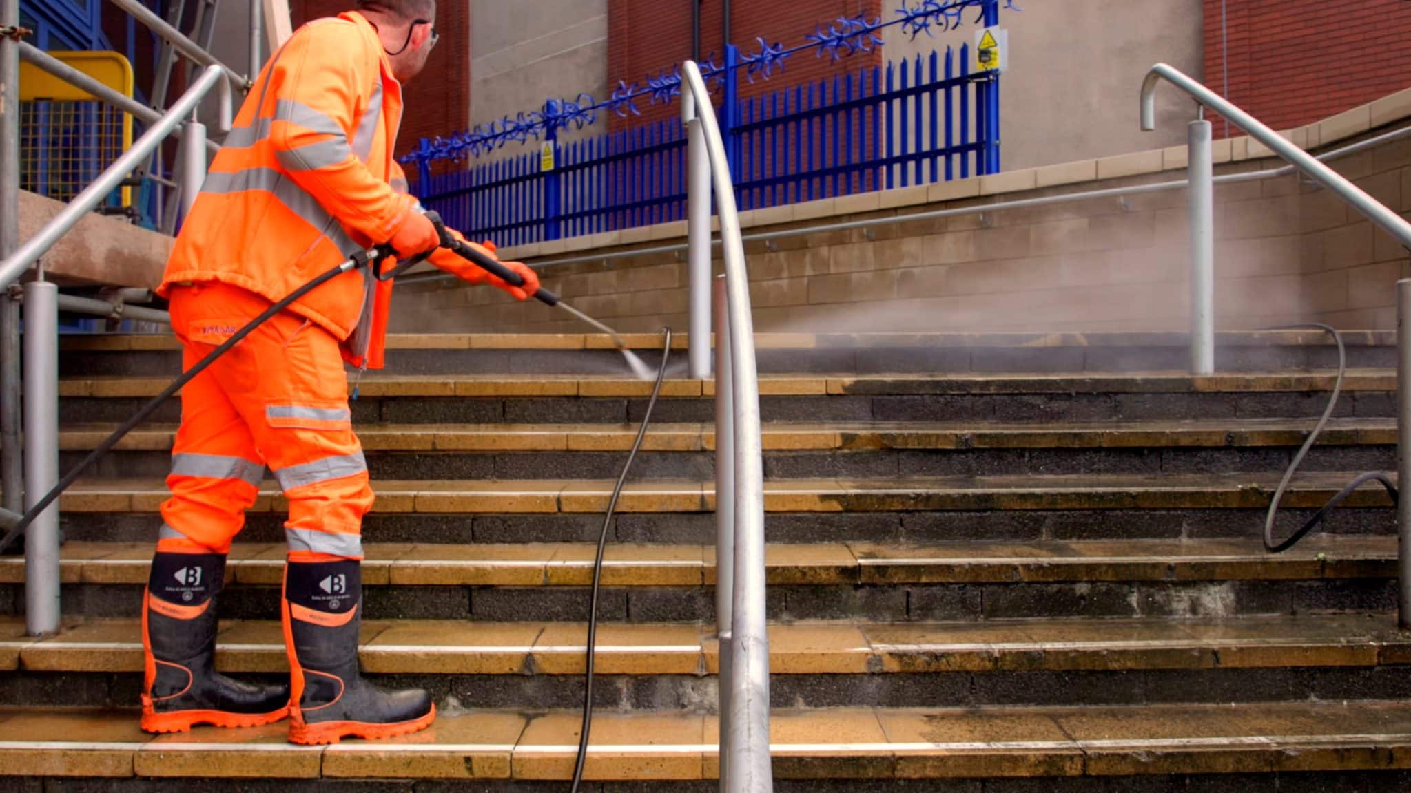 A person wearing an orange high-visibility suit, black rubber boots with orange soles, and safety glasses is using a pressure washer to clean a set of outdoor concrete and brick stairs with metal handrails. Water spray and steam are visible as they work. In the background, there is a blue metal fence with sharp spikes and a brick building.