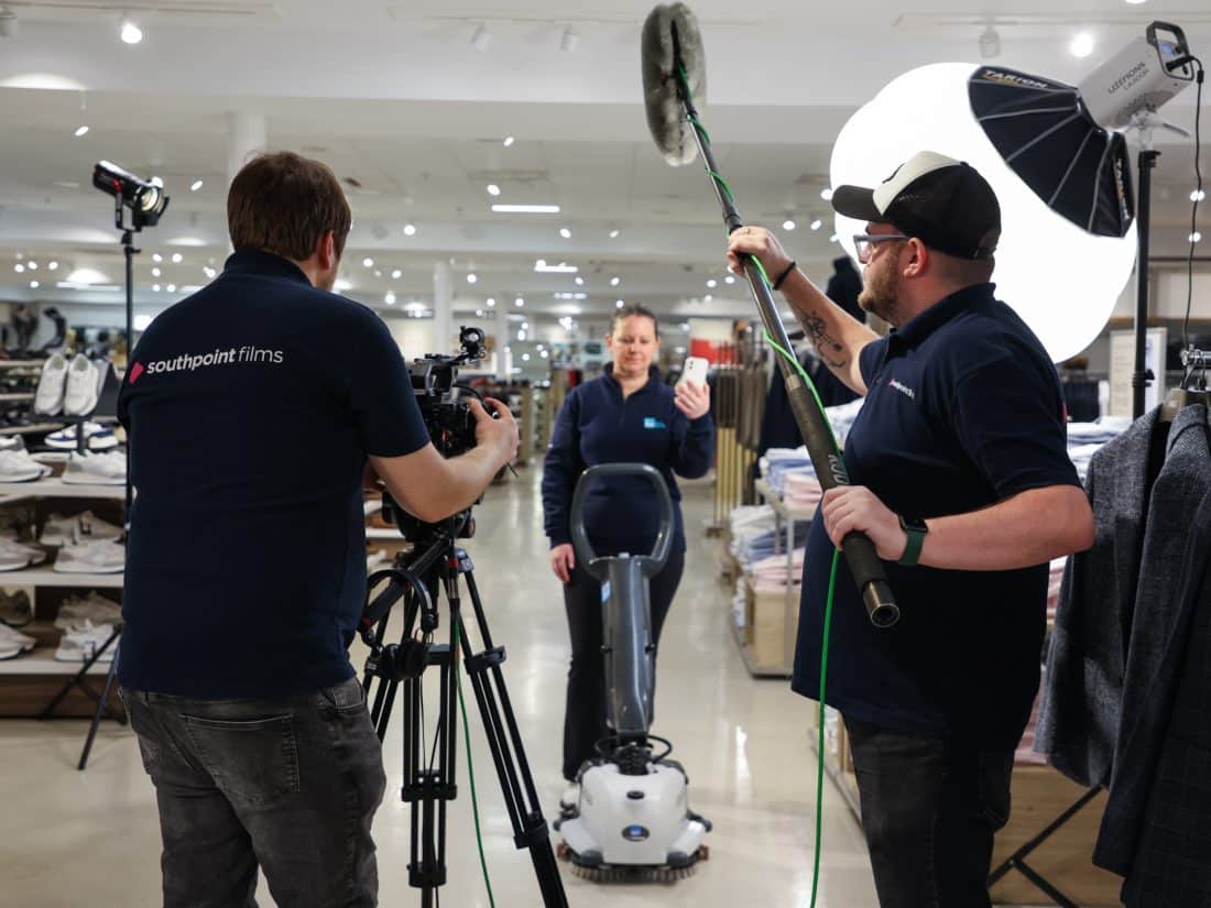 Three people are filming in a brightly lit retail store at M&S Hedge End, Southampton. A camera operator wearing a dark blue shirt with the word "southpoint films" on the back films a person operating a floor scrubbing machine in the centre. A sound operator on the right holds a boom microphone. Lighting equipment is set up to the right of the sound operator.