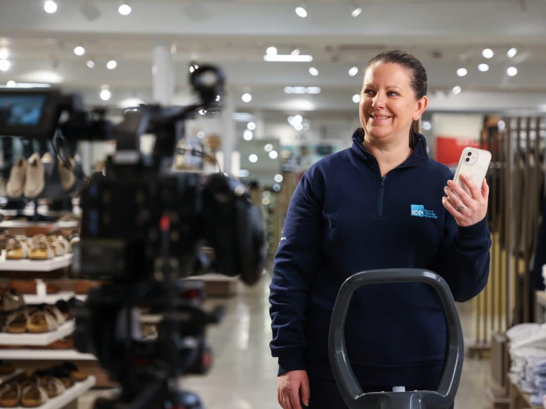 Woman in a dark blue, branded half-zip uniform stands in a brightly lit retail store holding a smartphone, looking toward a video camera in the foreground.