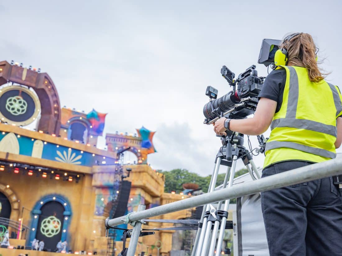 A camera operator wearing a high-visibility vest and headphones films a large, ornate outdoor festival stage under an overcast sky.