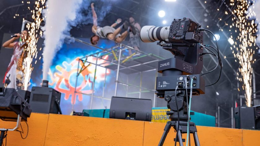 A professional video camera on a tripod is set up on an orange stage, capturing the action of a live performance. In the background, a person performs a mid-air backflip above a metal structure, while sparks shoot up from pyrotechnics on both sides of the stage.