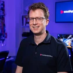 A person with brown hair and glasses wearing a dark polo shirt with the Southpoint Films logo, standing in a media production studio with blue ambient lighting and video equipment.