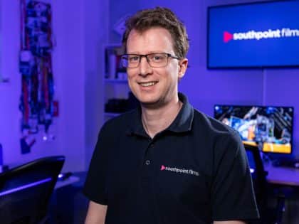 A person with brown hair and glasses wearing a dark polo shirt with the Southpoint Films logo, standing in a media production studio with blue ambient lighting and video equipment.
