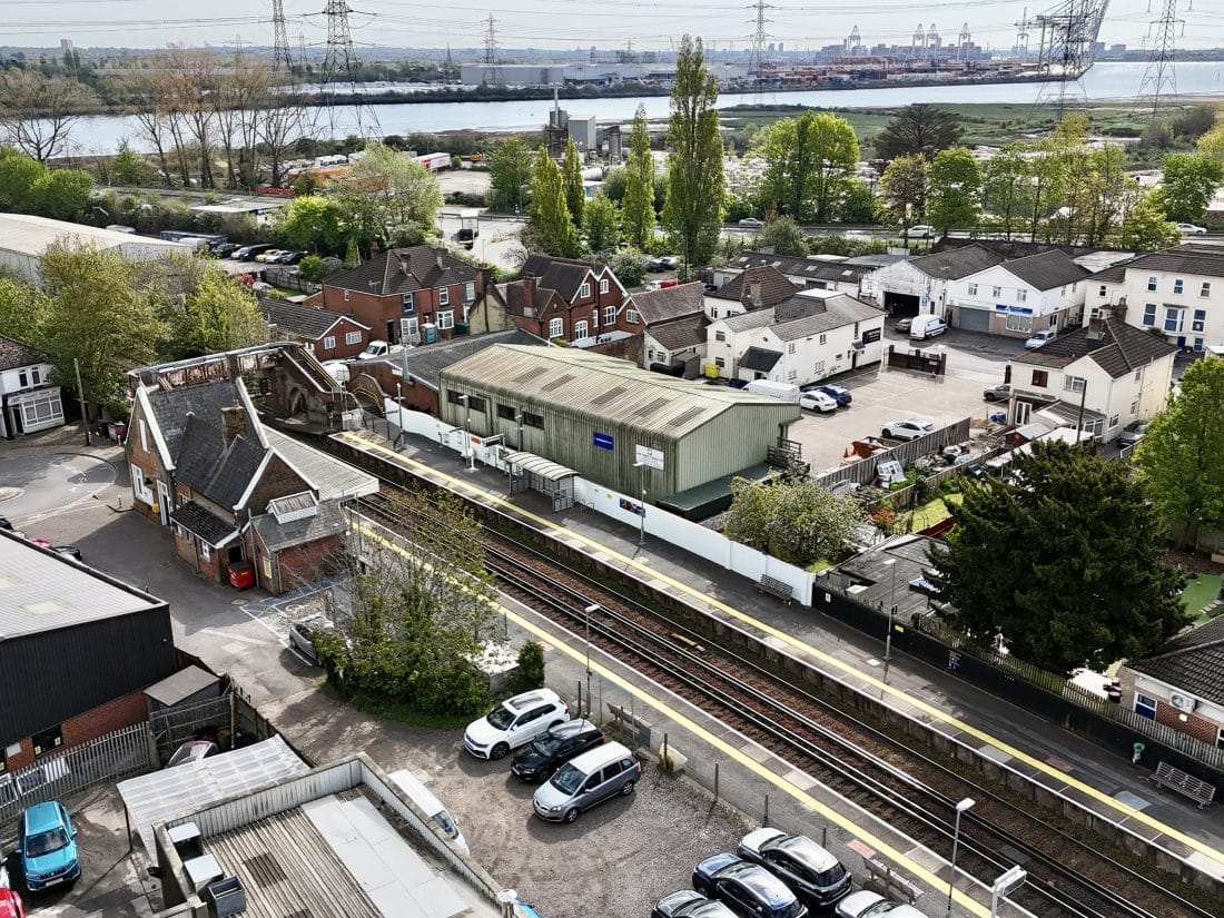 An aerial view of a small railway station featuring two tracks, a brick station building, and a pedestrian footbridge, surrounded by houses and industrial units with a river and shipping cranes in the distance.