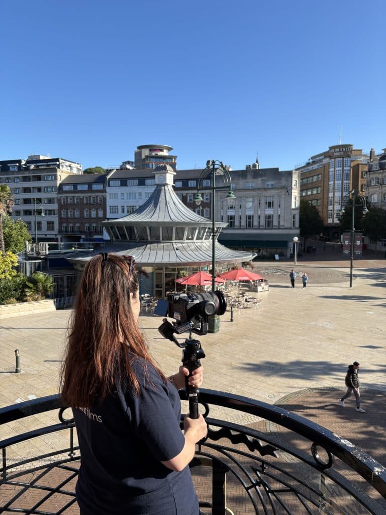 A person with long reddish-brown hair uses a camera mounted on a gimbal to film a sunny Bournemouth town centre, featuring a distinctive circular building with a tiered roof.