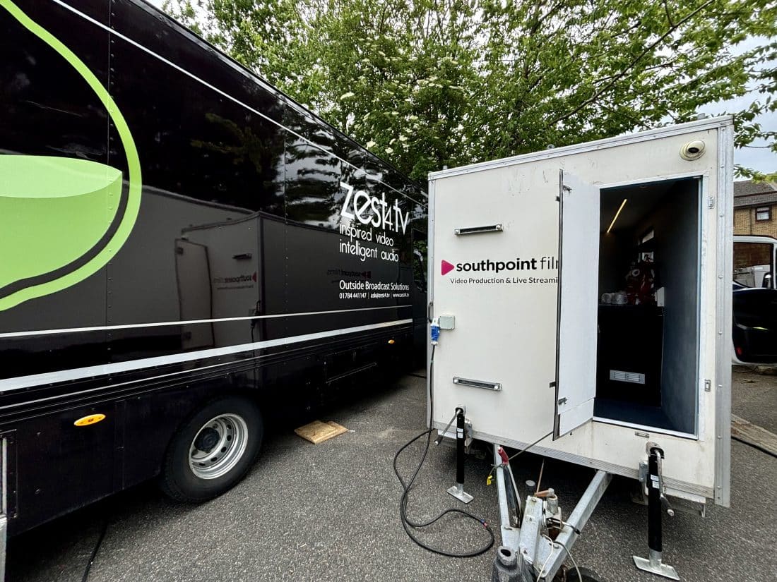 A large black outside broadcast truck with zest4.tv branding parked next to a white Southpoint Films trailer with its door open. The vehicles are positioned outdoors on pavement against a backdrop of green trees.